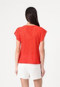 Top corto rosso a maniche corte con un design a occhiello che presenta forme a diamante. Il materiale è leggero, abbinato a pantaloncini bianchi per un look casual.