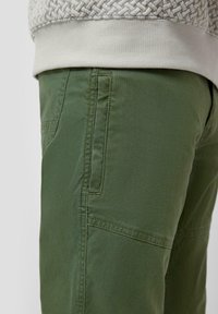 Zbliżenie zielonych spodni z widoczną boczną kieszenią i detalami szwów, w połączeniu z beżowym, teksturowanym karczkiem swetra.