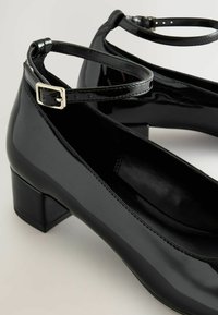 Schwarze lacklederne Blockabsatzschuhe mit Knöchelriemen und silbernen Schnallen, die eine glatte, glänzende Textur und ein rundes Zehendesign aufweisen.