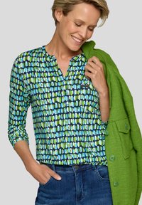 Mehrfarbige Bluse mit einem geometrischen Muster in Blau- und Grüntönen, mit einem V-Ausschnitt und langen Ärmeln, kombiniert mit einer grünen Jacke.