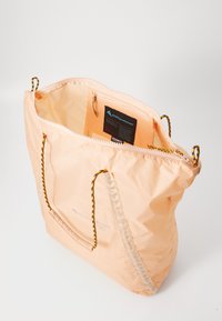 Perzikkleurige totebag van lichtgewicht stof met een ritssluiting aan de bovenkant, geweven handvatten en een binnenlabel met merk- en materiaalinformatie.