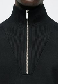 Μαύρο πουλόβερ με ψηλή λαιμόκοψη, διαθέτει υφή ριμπ, κλείσιμο με φερμουάρ μπροστά και σχέδιο σε σχήμα V στον γιακά. Ομαλό φινίρισμα υφάσματος.