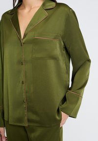 Sukienka z satyny w oliwkowym kolorze z kołnierzykiem, kieszonką z przodu i kontrastowym wykończeniem na krawędziach. Długie rękawy i zapięcie na guziki.