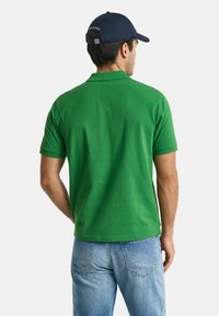 Groene katoenen polo met korte mouwen, gladde textuur, achterkant te zien met een eenvoudige uitstraling en een donkerblauwe pet. Lichte spijkerbroek zichtbaar.