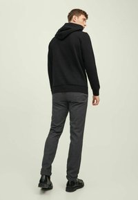 Čierna mikina s kapucňou spárovaná s tmavošedými slim-fit nohavicami a čiernymi topánkami, zobrazená zozadu na svetlozelenej pozadí.