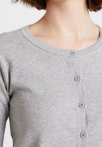 Harmaa neuletakki, joka on valmistettu pehmeästä kankaasta ja jossa on pyöreä pääntie. Edessä on napitus ja ribbineulekoristukset kauluksessa ja hihoissa.