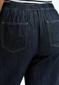 Jean en denim foncé avec une taille élastique, doté de deux poches arrière avec des contours cousus et une finition lisse et texturée.