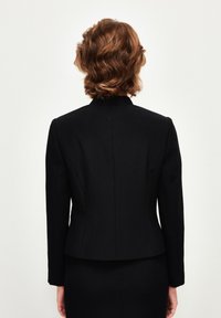 Czarna dopasowana marynarka z wyraźnymi ramionami i wysokim kołnierzem. Gładki materiał, długie rękawy i detale szwu z tyłu. Nosić na dopasowanej spódnicy.