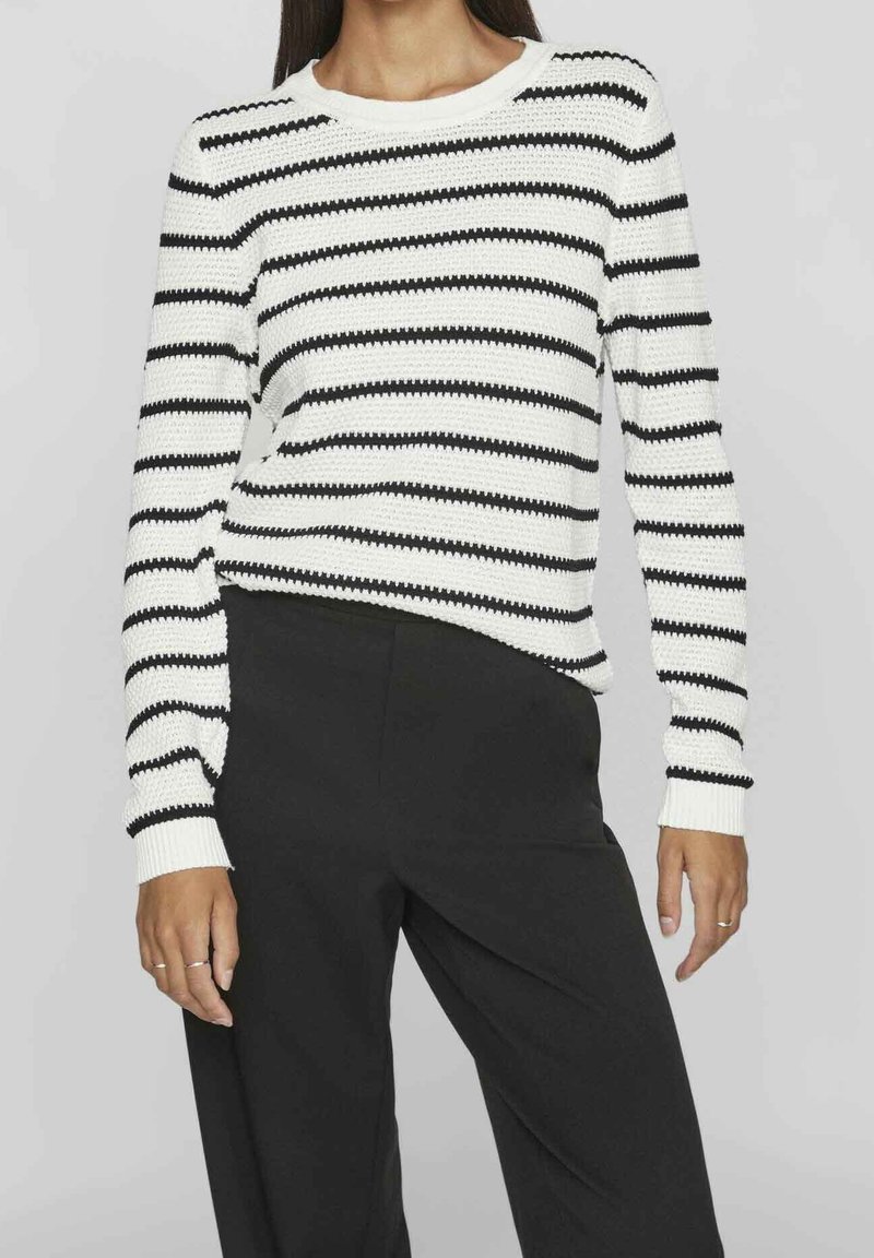 Femme portant un pull blanc à manches longues avec des rayures horizontales noires et un pantalon noir taille haute, debout devant un fond gris clair.