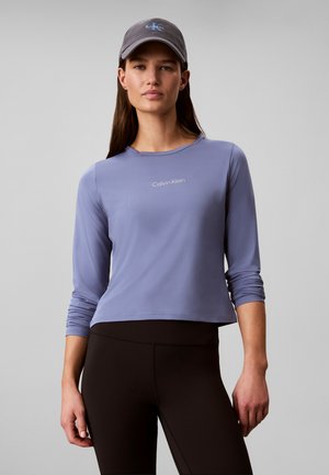 Kvinde iført en grå Calvin Klein kasket, en langærmet lavendel top med logo og sort højtaljede leggings mod en ensfarvet baggrund.