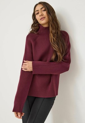 Burgundy turtleneck sweater with ribbed texture and long sleeves, featuring a relaxed fit and side slits, paired with black leggings.