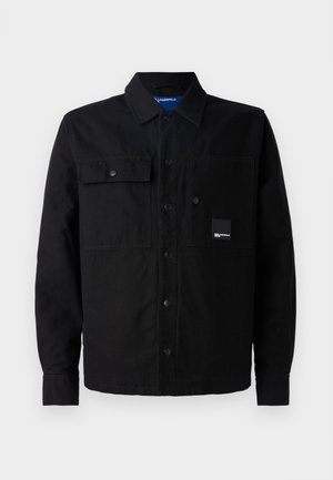 Black button-up shirt made of thick fabric, featuring two front pockets, snaps, and a logo patch on the left pocket. Classic design and fit.