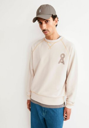 Mann trägt beige Sweatshirt mit Logo auf der Brust, graue Kappe mit Logo und blaue Jeans, steht vor weißem Hintergrund.