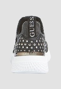 Talons de baskets Guess en noir et blanc avec des clous métalliques et le logo sur l'onglet de traction, ainsi qu'un accent métallique au-dessus de la semelle blanche texturée.
