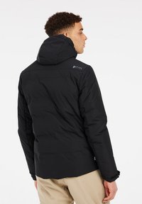 Chaqueta acolchada negra con capucha, que presenta una textura suave y un detalle de marca en la parte posterior, diseñada para ofrecer calidez y comodidad.