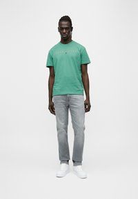 Πράσινο βαμβακερό μπλουζάκι με το κείμενο "TOMMY JEANS", συνδυασμένο με ανοιχτό γκρι τζιν παντελόνι και λευκά αθλητικά παπούτσια, στέκεται μπροστά σε απλό λευκό φόντο.