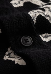 Crna pletena tkanina s blijedim uzorkom, s istaknutim crnim gumbom s četiri rupe, što stvara teksturirani izgled.