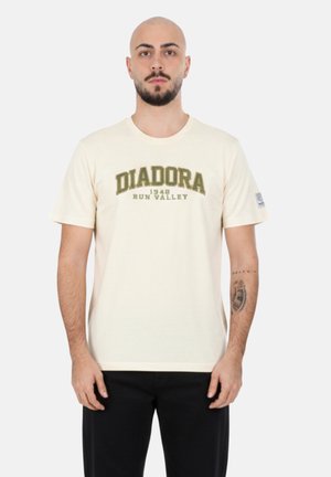 Uomo con barba e testa calva che indossa una maglietta beige Diadora Run Valley 1948 e pantaloni neri, in piedi contro uno sfondo bianco uniforme.