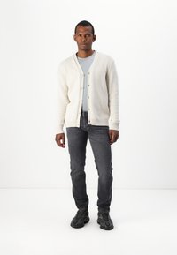 Cárdigan de punto color crema claro con cuello en V, cierre de botones y puños acanalados, combinado con una camiseta gris y jeans oscuros, sobre un fondo blanco.