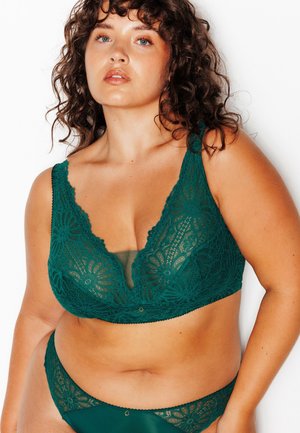 Femme aux cheveux bouclés portant un soutien-gorge et une culotte en dentelle vert foncé assortis, posant devant un fond blanc uni.