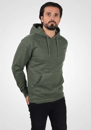 Groene hoodie met een trekkoordkap, voorzak en geribbelde manchetten. Gedragen met zwarte jeans, tegen een lichte vervagende achtergrond.