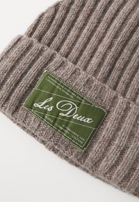 Beanie lavorato a maglia di colore marrone chiaro con una texture a coste. Presenta un'etichetta verde con testo bianco. Adatto per l'abbigliamento invernale.