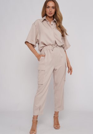 Kobieta ubrana w beżową, krótką koszulę zapinaną na guziki, wpuszczoną w beżowe, wysokie spodnie typu cargo z kieszeniami, stoi z jedną ręką w kieszeni.