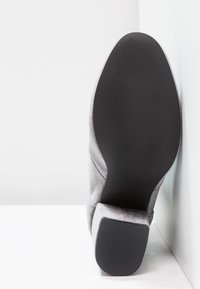 Semelle d'une botte à talon haut en daim gris avec une semelle extérieure en caoutchouc noir texturé et un talon bloc, positionnée debout contre un coin blanc.