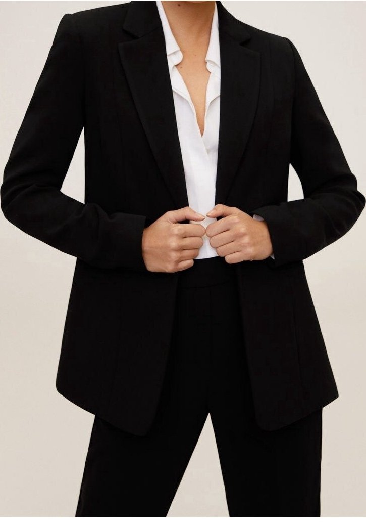 Personne portant un blazer noir sur une blouse blanche, les mains ajustant le blazer près de la taille, sur un fond clair uni.