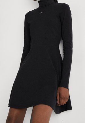 Femme portant une robe noire à manches longues avec col roulé, cintrée à la taille, s'étendant jusqu'à mi-cuisse.
