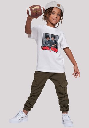 Das weiße T-Shirt zeigt ein Grafik von Laurel und Hardy. Das Kind trägt olivgrüne Cargo-Hosen und weiße Turnschuhe mit hellgrauen Akzenten und hält einen braunen Fußball.