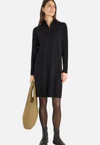 Abito nero in maglia con colletto a zip, dettagli a costine e maniche lunghe. Abbinato a una borsa beige e stivaletti neri con lacci.