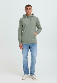 Sudadera gris-verde con un bolsillo frontal, capucha con cordón y puños de canalé, combinada con jeans de color azul claro y zapatillas blancas.