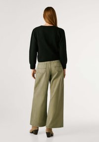 Melna adīta džemperis ar izteiksmīgām piedurknēm, saskaņots ar plašām olīvu zaļām biksēm un blokveida papēžiem, skatīts no aizmugures.