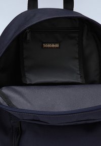 Interior de mochila de tela azul marino con un bolsillo negro con cremallera, que cuenta con una etiqueta de logo. Superficie texturizada, compartimento principal espacioso, apertura en la parte superior.