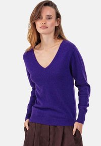 V-Ausschnitt lila Pullover aus weichem Strickstoff, mit langen Ärmeln und gerippten Bündchen an Ärmeln und Saum. Kombiniert mit einem braunen Rock.