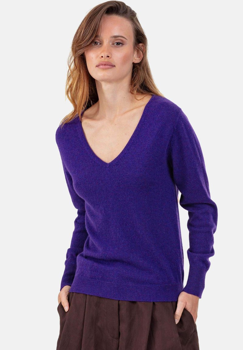 V-Ausschnitt lila Pullover aus weichem Strickstoff, mit langen Ärmeln und gerippten Bündchen an Ärmeln und Saum. Kombiniert mit einem braunen Rock.