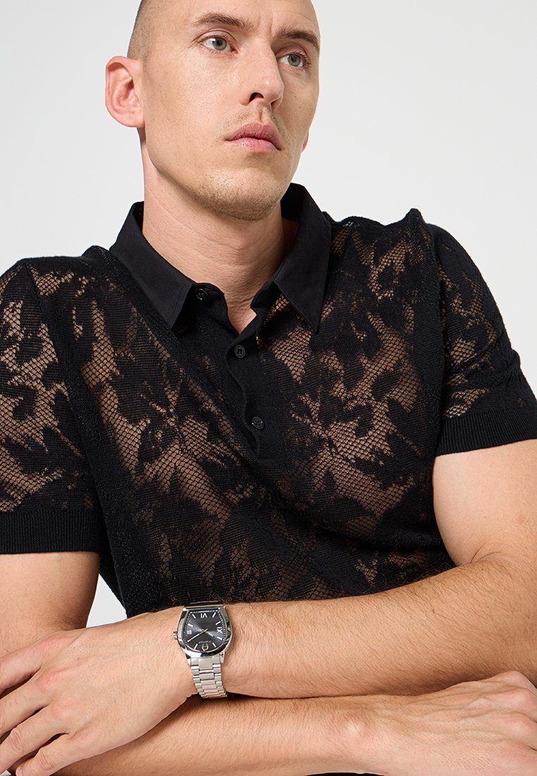 Schwarzes Poloshirt mit einem durchsichtigen Spitzenmuster und einem festen Kragen. Das Model trägt eine silberne Uhr mit rechteckigem Ziffernblatt an einem Metallarmband.