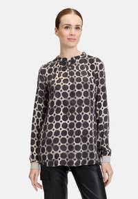 Long-sleeve blouse in cream with large black circles, featuring a pleated neckline. Cuffs have a glittery accent. Paired with black pants.