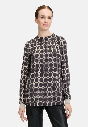 Langärmlige Bluse in Creme mit großen schwarzen Kreisen, ausgestattet mit einem plissierten Ausschnitt. Die Manschetten haben einen glänzenden Akzent. Kombiniert mit schwarzen Hosen.