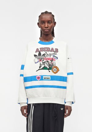 Person trägt weißen Adidas-Sweatshirt mit blauen Streifen und sportlichem Motiv, kombiniert mit schwarzen Adidas-Trainingshosen mit weißen Streifen.