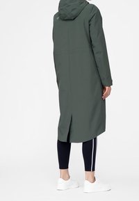 Lang, hætteklædt regnfrakke i mørkegrønt stof med en bærestykkeventilation bagpå og et enkelt design, kombineret med sorte leggings og hvide sneakers.