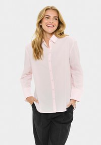 Femme souriante avec de longs cheveux blonds portant une chemise à boutons rose clair et un pantalon noir, les mains dans les poches, sur fond uni.