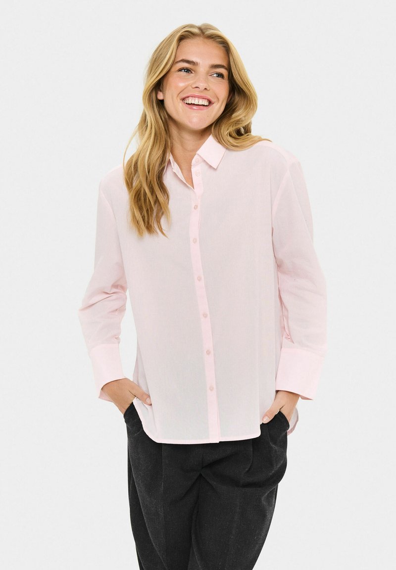 Femme souriante avec de longs cheveux blonds portant une chemise à boutons rose clair et un pantalon noir, les mains dans les poches, sur fond uni.