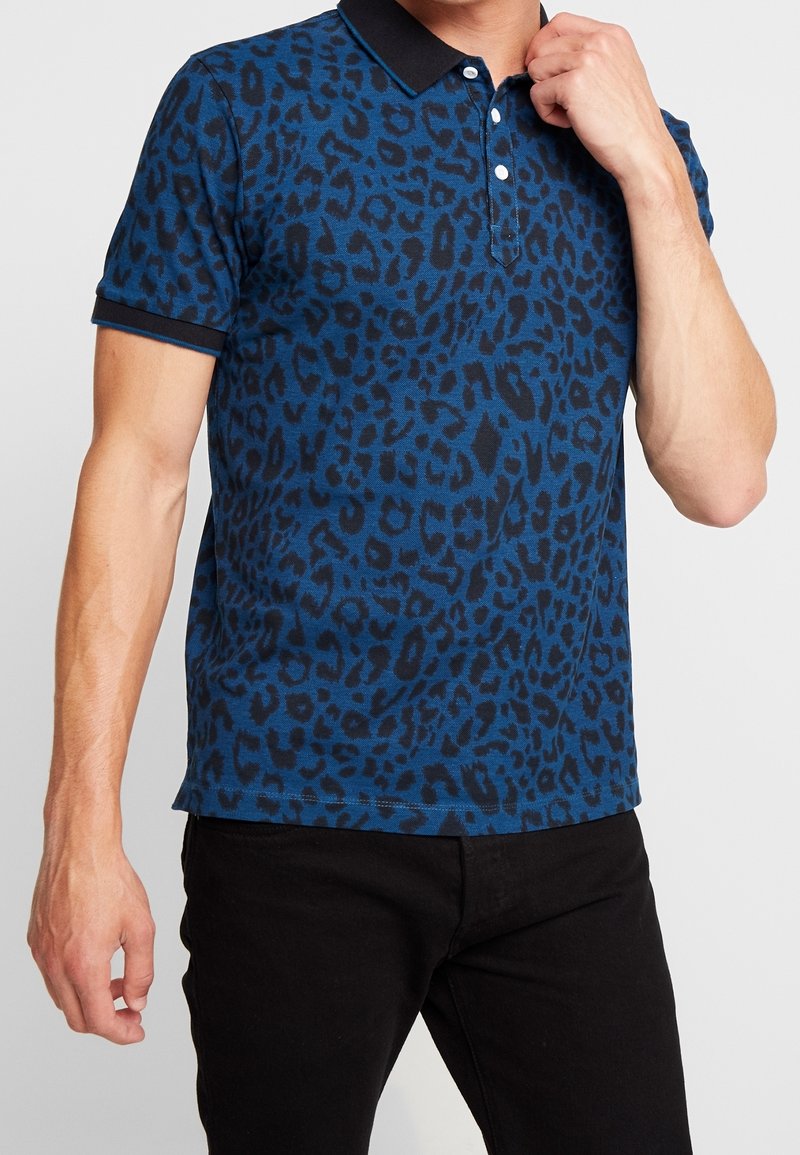 Homme portant un polo bleu à manches courtes avec imprimé léopard noir et pantalon noir, main près du col.