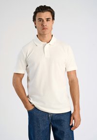 Kortärmad creamfärgad pikétröja i ett strukturerat tyg med krage och tre-knappsprydd öppning, kombinerad med blå jeans i denim.