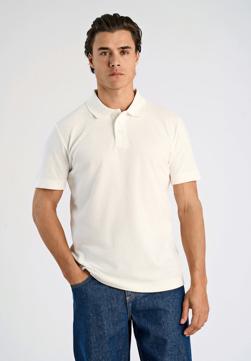 Kortärmad creamfärgad pikétröja i ett strukturerat tyg med krage och tre-knappsprydd öppning, kombinerad med blå jeans i denim.