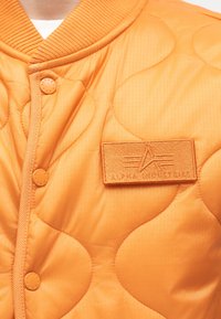 Veste matelassée orange avec col côtelé, boutons-pression et écusson rectangulaire du logo Alpha Industries sur la poitrine gauche.