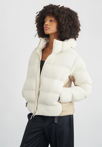 Kobieta o kręconych włosach, ubrana w białą pikowaną kurtkę z kapturem i czarne spodnie, stojąca z rękami w kieszeniach na jednolitym białym tle.