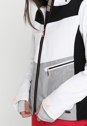 Femme portant une veste zippée blanche, grise et noire avec des trous pour les pouces, tenant l'ourlet inférieur au-dessus d'un pantalon rouge, debout devant un fond uni.